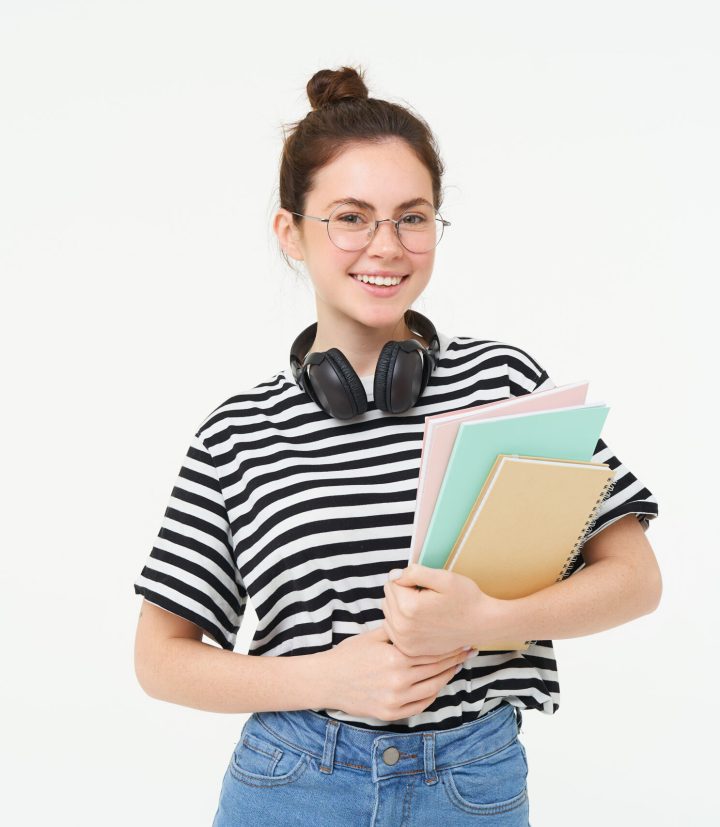 Education concept. Smiling brunette girl, student in casual clothes, holds her books, study material, wears headphones over neck, looks confident and happy, isolated over white background Education concept. Smiling brunette girl, student in casual clothes, holds her books, study material, wears headphones over neck, looks confident and happy, isolated over white background.