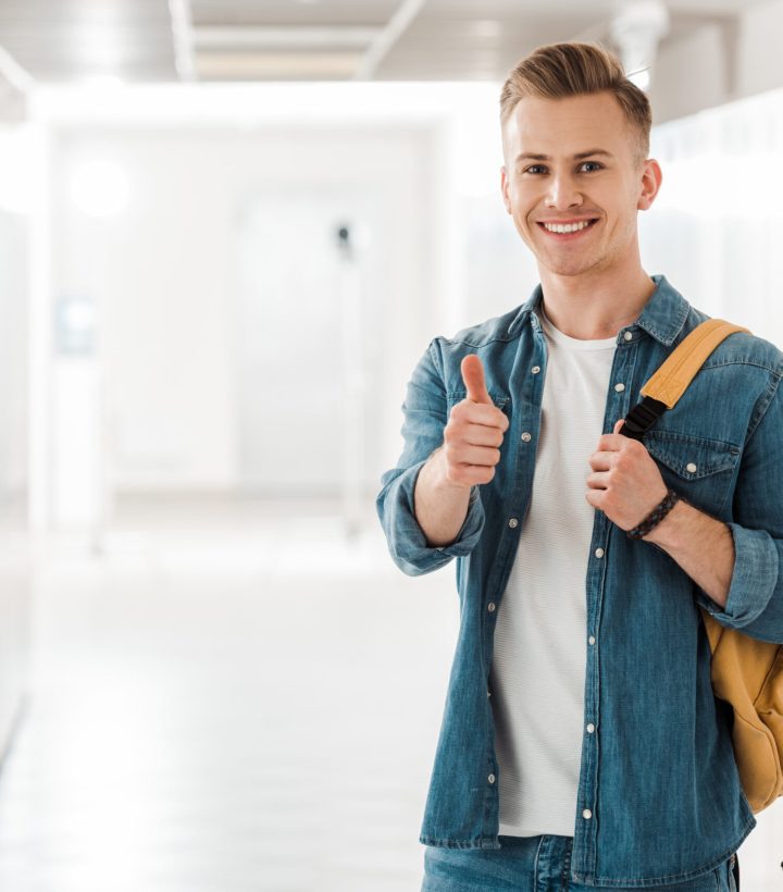 student with backpack showing thumb up in corridor in university student with backpack showing thumb up in corridor in university